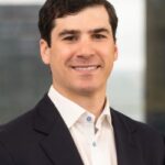 Professional headshot of a smiling man in a dark blazer and white shirt, with a blurred office background.