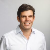 Head-and-shoulders portrait of a smiling man in a white button-down shirt against a light gray background.