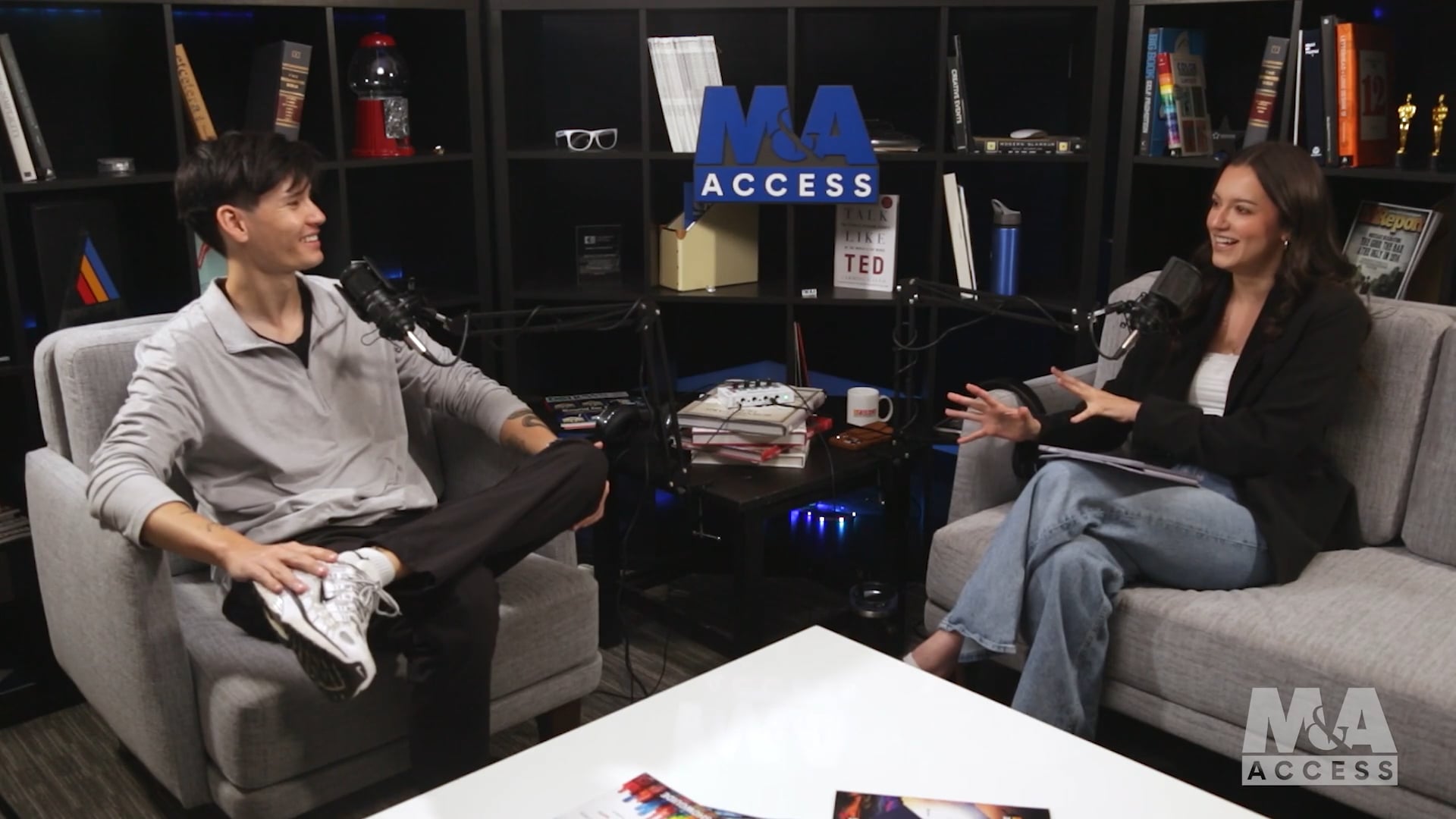 Two podcast hosts sit on gray couches facing each other, speaking into microphones with a bookshelf backdrop and the M&A ACCESS logo visible behind them.