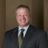 John Cohan Professional headshot of a smiling man in a dark suit, light blue shirt, and gold tie, against a brown backdrop.